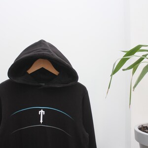 May include: Black hooded sweatshirt with a white and blue graphic design. The sweatshirt has a drawstring hood and a kangaroo pocket. The graphic features a stylized letter "T" with a curved line above it. The sweatshirt is hanging on a wooden hanger.