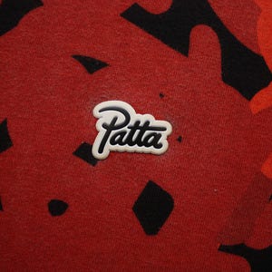 Puede incluir: Primer plano de una tela roja con un patr&oacute;n de camuflaje negro y rojo. El logotipo "Patta" blanco y negro est&aacute; centrado en la tela. La tela tiene una apariencia texturizada.