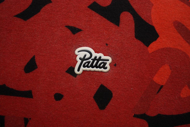 Puede incluir: Primer plano de una tela roja con un patr&oacute;n de camuflaje negro y rojo. El logotipo "Patta" blanco y negro est&aacute; centrado en la tela. La tela tiene una apariencia texturizada.