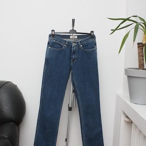 May include: Dark blue denim jeans hanging on a black hanger. The jeans have a classic five-pocket design and a straight leg cut. The brand name "Acne" is visible on the front pocket. The jeans are displayed against a white wall.