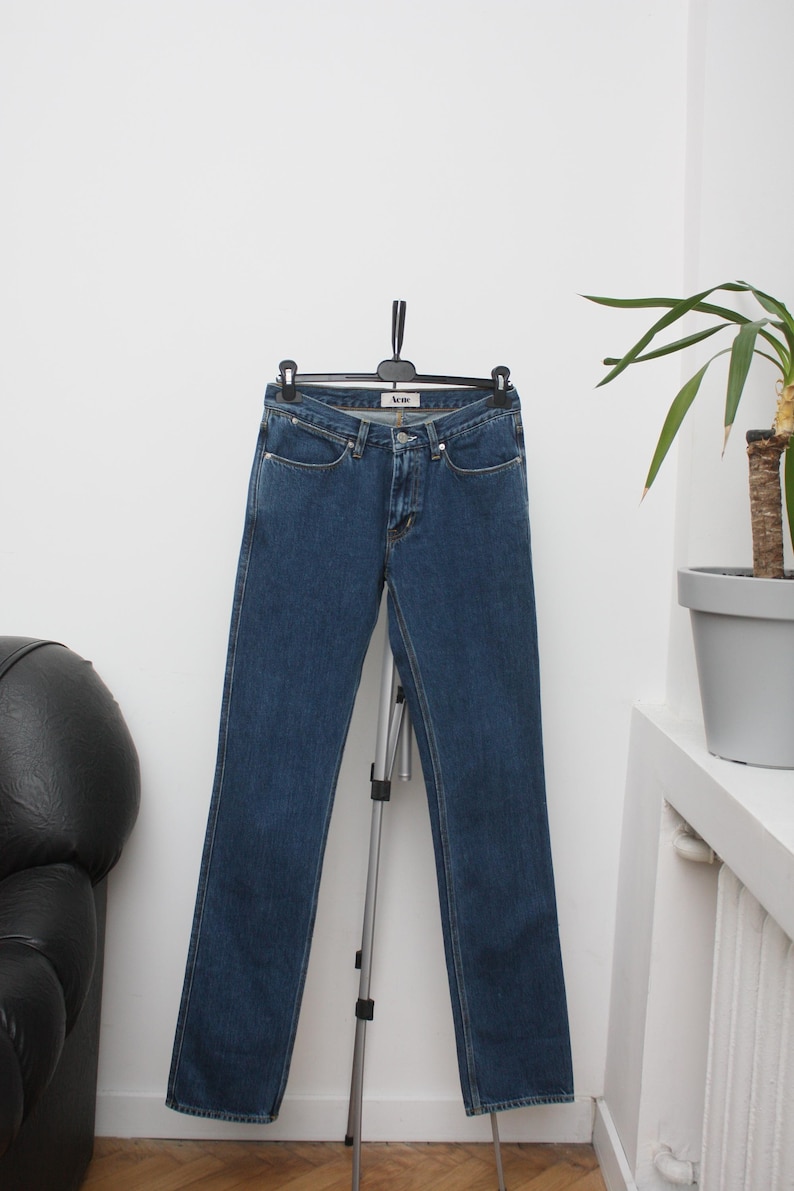 May include: Dark blue denim jeans hanging on a black hanger. The jeans have a classic five-pocket design and a straight leg cut. The brand name "Acne" is visible on the front pocket. The jeans are displayed against a white wall.