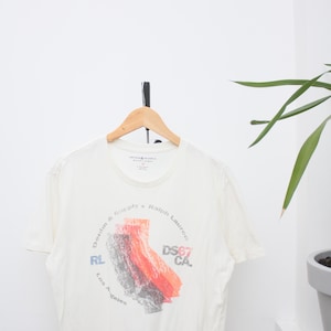 May include: Cream t-shirt with a graphic of the California state outline in red, grey, and black. The shirt features the text "Denim & Supply Ralph Lauren" and "DS67 CA". The t-shirt is hanging on a wooden hanger.