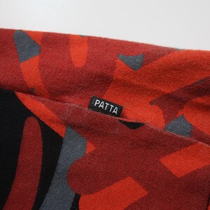 Puede incluir: Primer plano de una tela roja con un patr&oacute;n abstracto negro y gris. La tela tiene una etiqueta que dice "PATTA". El material parece ser una tela de punto o jersey, con costuras visibles.