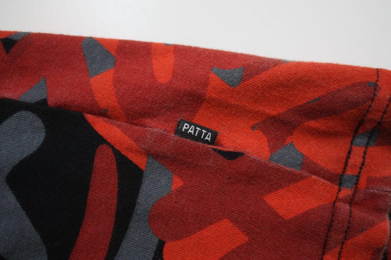 Puede incluir: Primer plano de una tela roja con un patr&oacute;n abstracto negro y gris. La tela tiene una etiqueta que dice "PATTA". El material parece ser una tela de punto o jersey, con costuras visibles.