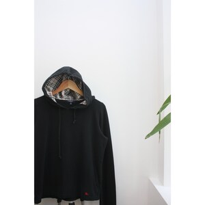 May include: Black hooded sweatshirt with a plaid lining inside the hood. The garment is displayed on a wooden hanger. A small red logo is visible near the hem. The background is white.