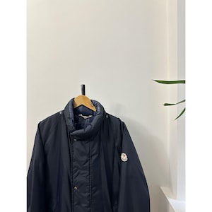 Puede incluir: Chaqueta acolchada azul marino con cuello alto y un pequeño parche con el logo en la manga. La chaqueta está colgada en una percha de madera contra una pared blanca. La chaqueta tiene un diseño acolchado y cierre de botones.