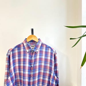 Peut inclure: Chemise &agrave; carreaux &agrave; manches longues dans les tons de violet, rose, blanc et bleu, suspendue &agrave; un cintre en bois. La chemise a un col boutonn&eacute; et la marque "Acne" est visible sur le col.