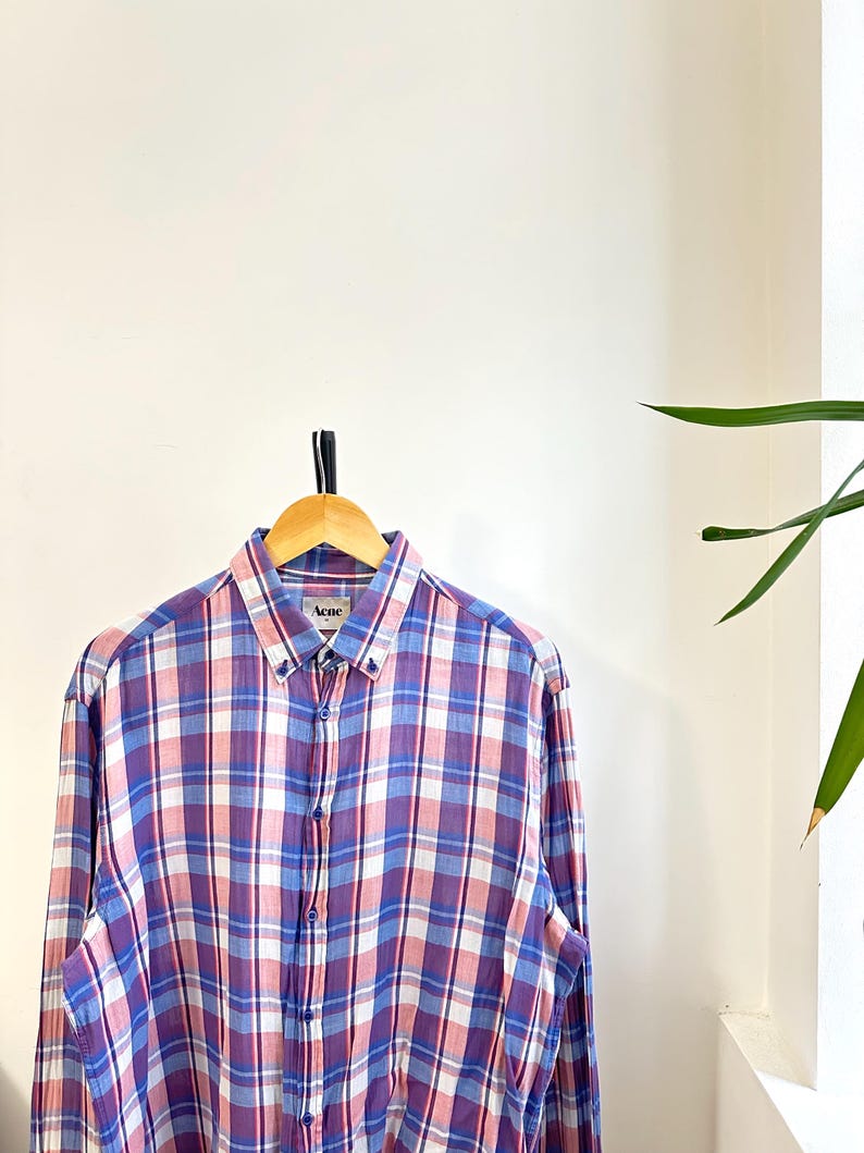 Peut inclure: Chemise &agrave; carreaux &agrave; manches longues dans les tons de violet, rose, blanc et bleu, suspendue &agrave; un cintre en bois. La chemise a un col boutonn&eacute; et la marque "Acne" est visible sur le col.