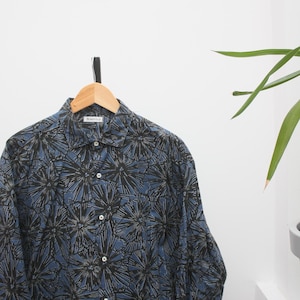 May include: A button-down shirt with a blue background and a black and gray floral pattern. The shirt is hanging on a wooden hanger against a white wall. The label reads "Honeyville".