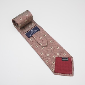 Puede incluir: Una corbata Givenchy con estampado rojo y blanco. La corbata está enrollada en la parte superior y colocada sobre una superficie blanca. Tiene un patrón repetitivo de pequeños círculos blancos con un centro rojo oscuro. Se ve una etiqueta azul marino con el logotipo de Givenchy.