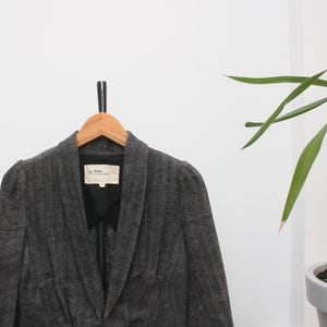 May include: A gray herringbone blazer with a notched lapel and a single-button closure. The blazer is hanging on a wooden hanger against a white wall. The blazer has two front pockets and a label inside the collar.