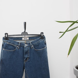 May include: Dark blue denim jeans hanging on a black hanger. The jeans have a classic five-pocket design and a straight leg cut. The brand name "Acne" is visible on the waistband. The jeans are displayed against a white background.