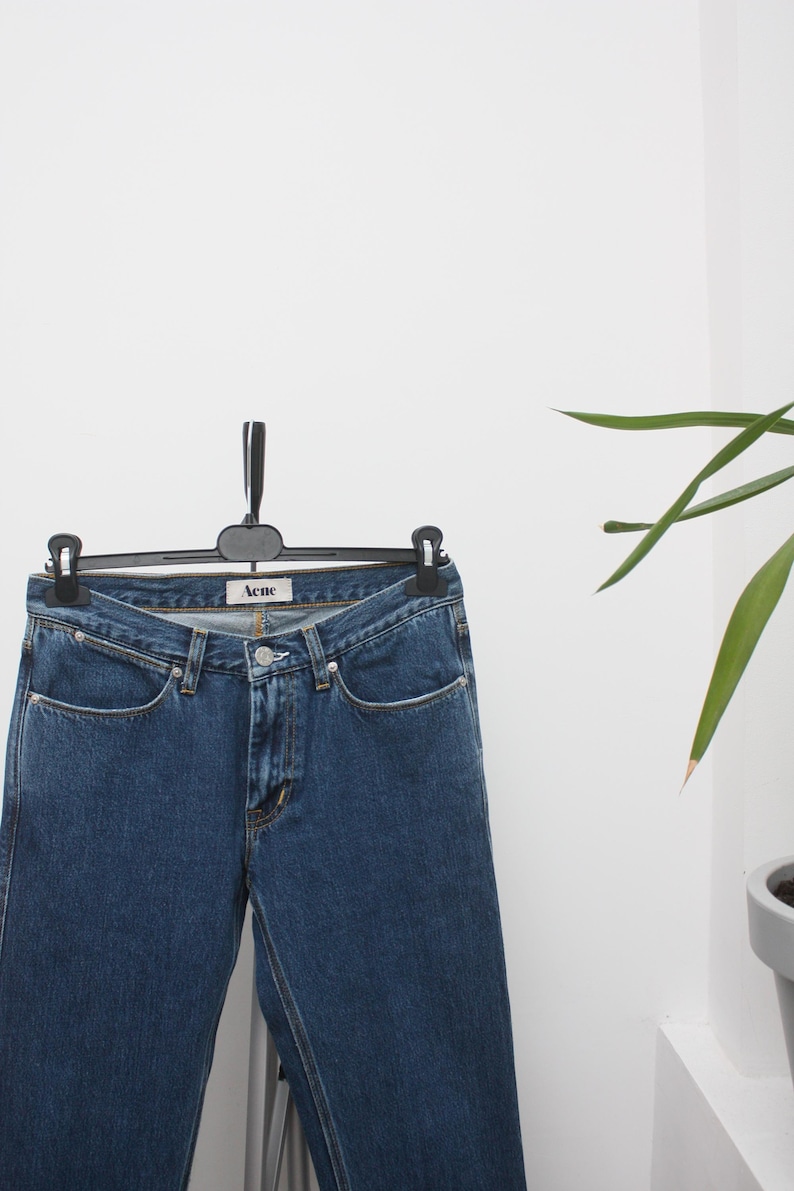 May include: Dark blue denim jeans hanging on a black hanger. The jeans have a classic five-pocket design and a straight leg cut. The brand name "Acne" is visible on the waistband. The jeans are displayed against a white background.
