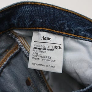 May include: Close-up of blue denim jeans with a visible label. The label reads "Acne" and includes size information, "30/34". The jeans are made of cotton and suede. The stitching is visible.