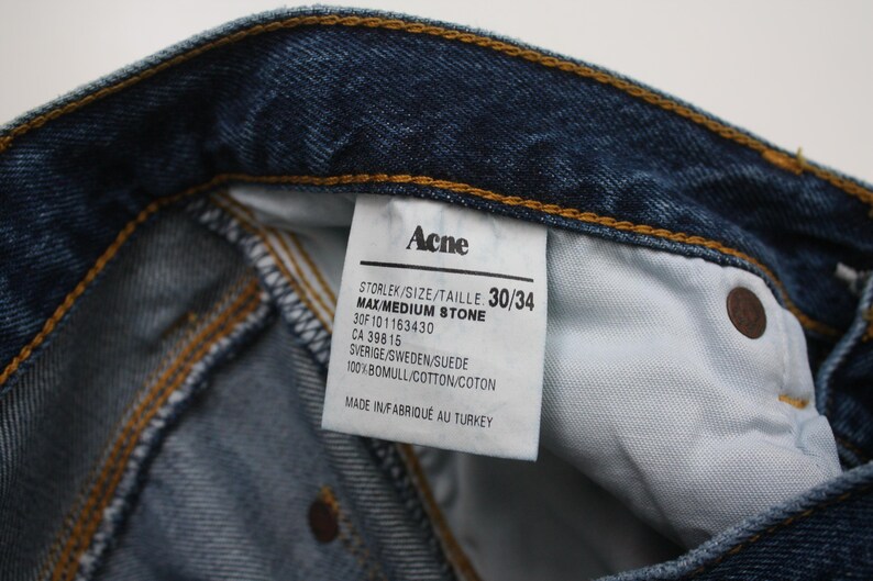 May include: Close-up of blue denim jeans with a visible label. The label reads "Acne" and includes size information, "30/34". The jeans are made of cotton and suede. The stitching is visible.
