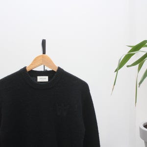 May include: A black crew neck sweater with a textured knit. The sweater features a small, tonal embroidered logo on the chest and a label at the neckline. It hangs on a wooden hanger against a white wall.