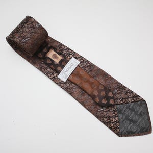 May include: A brown silk tie with a repeating floral pattern and a solid brown center. The tie has a label that reads "VERSACE". The tie is folded and lies on a white surface.