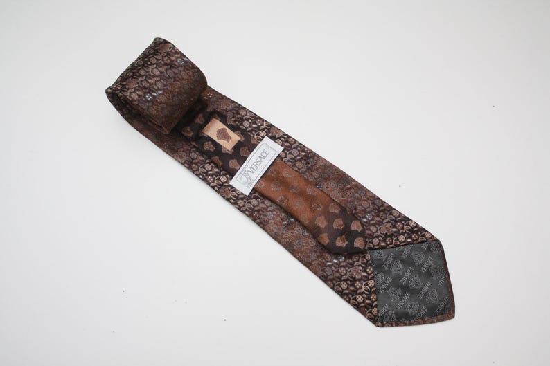 May include: A brown silk tie with a repeating floral pattern and a solid brown center. The tie has a label that reads "VERSACE". The tie is folded and lies on a white surface.