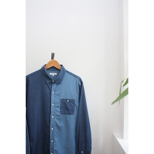 May include: A two-tone denim shirt in shades of blue hangs on a wooden hanger. The shirt features a button-down front, a chest pocket, and long sleeves. The color-blocked design creates a visual contrast, with one side a darker blue than the other.