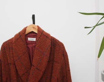 May include: A rust-colored, textured coat with a shawl collar and a matching belt. The coat features a subtle plaid pattern in navy blue and is hanging on a wooden hanger. The lining is a deep burgundy color.