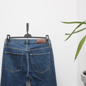 May include: Blue denim jeans hanging on a black hanger. The jeans have two back pockets and a brown leather patch with the brand name. The jeans are a medium wash and appear to be new.