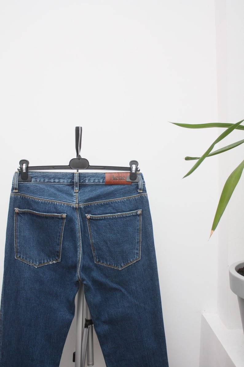 May include: Blue denim jeans hanging on a black hanger. The jeans have two back pockets and a brown leather patch with the brand name. The jeans are a medium wash and appear to be new.