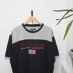 May include: A black and grey Chaps Ralph Lauren t-shirt with a small American flag. The shirt has short sleeves and a crew neck. The Chaps logo is embroidered in red.