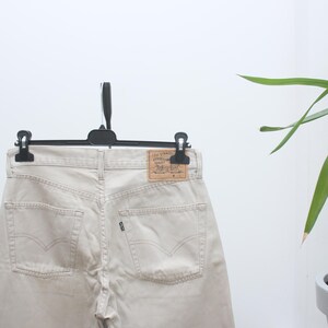 Puede incluir: Unos vaqueros Levi's 501 de color beige claro colgados de una percha negra. Los vaqueros tienen el clásico diseño de cinco bolsillos, bragueta con botones y parche de cuero. Los vaqueros son de tejido vaquero resistente.