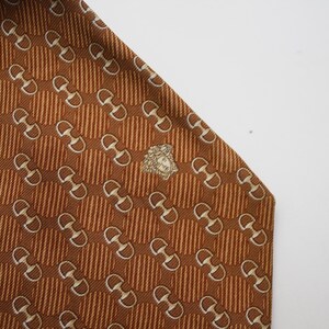 Op de afbeelding: Een bruine zijden stropdas met een herhalend patroon van witte paardenbitjes en verticale strepen. Een goudkleurig embleem bevindt zich in de buurt van de onderkant. De stropdas staat schuin tegen een witte achtergrond.