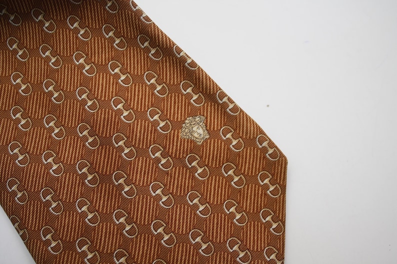 Op de afbeelding: Een bruine zijden stropdas met een herhalend patroon van witte paardenbitjes en verticale strepen. Een goudkleurig embleem bevindt zich in de buurt van de onderkant. De stropdas staat schuin tegen een witte achtergrond.