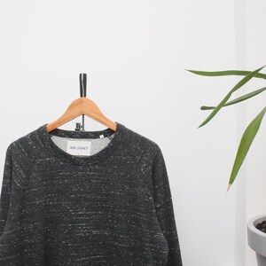 May include: A dark gray sweatshirt with white flecks hangs on a wooden hanger. The sweatshirt has a crew neck and long sleeves. The text "OUR LEGACY" is visible on the label. The background is white with a green plant.