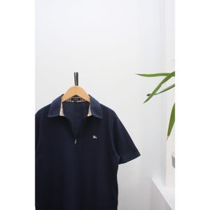 May include: A navy blue short-sleeved polo shirt with a zip-up collar. The collar and inner lining have a plaid pattern. A small emblem is visible on the chest. The shirt is hanging on a wooden hanger.