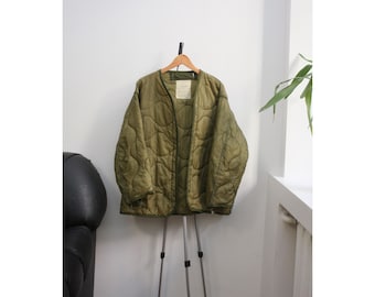 Puede incluir: Chaqueta acolchada verde oliva con un patrón de diamantes, colgada de una percha de madera. La chaqueta tiene una etiqueta en el pecho. Se exhibe en un trípode plateado contra una pared blanca.
