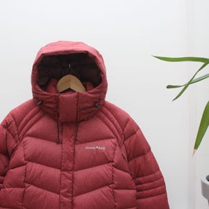 May include: A burgundy puffer jacket with a hood, hanging on a wooden hanger. The jacket has a quilted design and the brand name "mont bell" is visible on the chest. The lower portion of the jacket is a darker shade of burgundy.