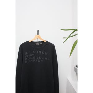 May include: Black crew neck sweatshirt with the text "R. Lauren 19-67 Polo Jeans Company" printed on the front. The sweatshirt is hanging on a wooden hanger against a white wall. The fabric has a waffle-like texture.