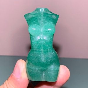 May include: A green gemstone sculpture of a female torso. The sculpture is carved from a translucent, green stone, with smooth, rounded forms. The figure is depicted without arms or legs, with a focus on the upper body.