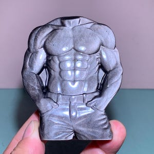 May include: A gray, sculpted torso figurine depicting a muscular physique. The figure is detailed with defined abs, chest muscles, and arms, and is wearing pants with a belt. The figurine is held by a hand.