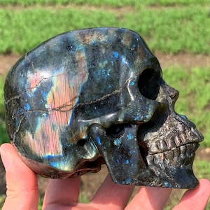 Puede incluir: Un cráneo tallado en piedra labradorita. El cráneo tiene un color base oscuro con destellos iridiscentes azules, verdes y rosas. Los detalles del cráneo, incluyendo las cuencas de los ojos y los dientes, están bien definidos.