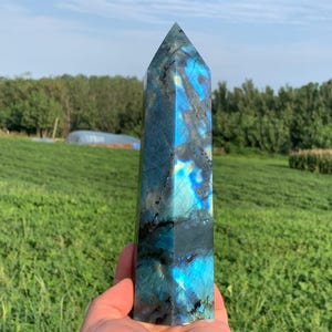 May include: A tall, rectangular labradorite crystal obelisk with a pointed top. The stone displays iridescent blue, teal, and black hues. The obelisk is held in front of a green field and trees.