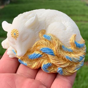 May include: A white fox figurine in a sleeping pose, with gold and blue accents on its tail. The fox has a gold embellishment on its forehead. The figurine is held in a hand against a blurred green background.