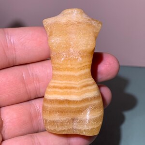 May include: A carved, yellow and brown stone figurine in the shape of a dressmaker's form. The figurine has a smooth, polished surface with horizontal bands of color. The form is a miniature sculpture, likely for display or collection.