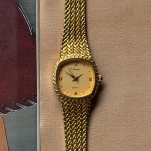 May include: A gold-tone wristwatch with a textured, woven-style metal band and a square-shaped case with rounded edges. The dial is gold with a fine cross-hatch pattern, featuring the text Pi Camin, Quartz, and Japan Movt, with small stone markers.