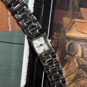 May include: A silver-toned Bulova wristwatch featuring a rectangular face with a white dial, silver hands, and a matching metallic link bracelet. The watch displays the Bulova brand name on the dial and includes a small date window at the three o'clock position.