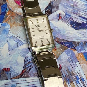 May include: A rectangular silver-tone Citizen watch with a white dial, featuring the text "xC" and "CITIZEN" on the face. The watch has a metallic link bracelet, silver hour markers, and three thin hands, presented in a clean, functional design.