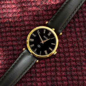 May include: A gold-toned Gucci watch with a black face and black leather band. The watch face features gold hands and Roman numeral hour markers. The watch band is black leather with gold-toned hardware.