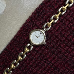 May include: A silver and gold-toned Alba Pingo wristwatch with a white face and a linked bracelet. The watch has a round face with black hands and hour markers. The bracelet alternates silver and gold-toned links. The watch is displayed on a burgundy knitted surface.