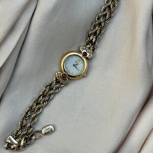May include: Gold-toned wristwatch with a white face and gold hands. The watch has a round face with a gold bezel and is adorned with small red gemstones. The watch band is a braided design.
