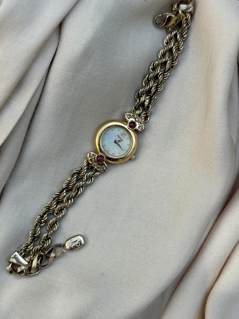 May include: Gold-toned wristwatch with a white face and gold hands. The watch has a round face with a gold bezel and is adorned with small red gemstones. The watch band is a braided design.