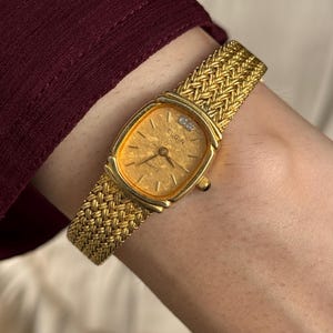 May include: A gold-toned Elgin quartz watch with a textured face and a small diamond accent. The watch has a woven gold band. The watch is displayed on a wrist.