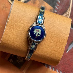 Puede incluir: Un reloj de pulsera rígida en tono plateado con una esfera redonda sobre un fondo azul intenso y una ilustración de una flor clara en el centro. La banda está decorada con paneles de esmalte azul con pequeños motivos florales blancos.